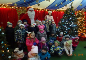 Dzieci pozują do zdjęcia ze Świetym Mikołajem i Śnieżynką. Po bokach stoją choinki.
